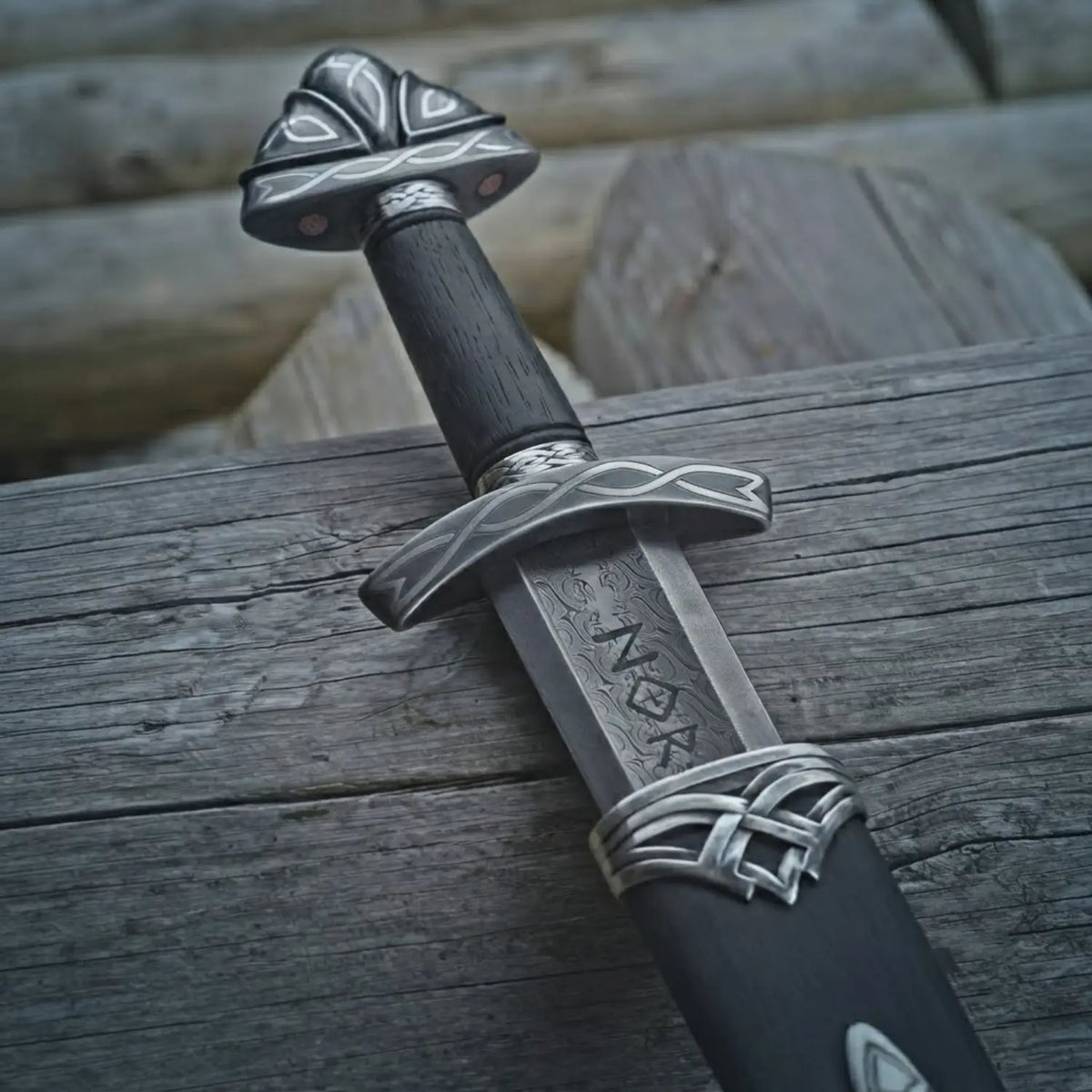 Hand-forged Viking Seax sword with Damascus steel blade, ornate silver fittings, and black wood handle resting on rustic wooden planks.