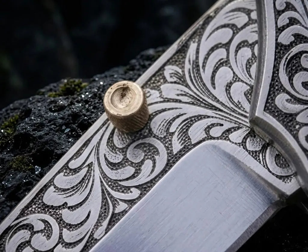 Extreme close-up macro view of intricate hand-engraved scrollwork pattern on steel blade bolster with solid brass thumb stud on custom pocket knife.