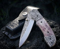 Handmade engraved Damascus-style folding pocket knife with intricate scrollwork, smooth natural bone handle, brass thumb stud, and black leather belt sheath displayed on volcanic rock.
