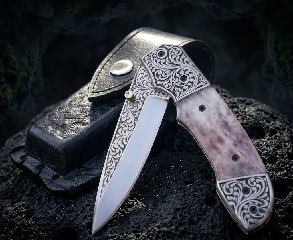 Handmade engraved Damascus-style folding pocket knife with intricate scrollwork, smooth natural bone handle, brass thumb stud, and black leather belt sheath displayed on volcanic rock.
